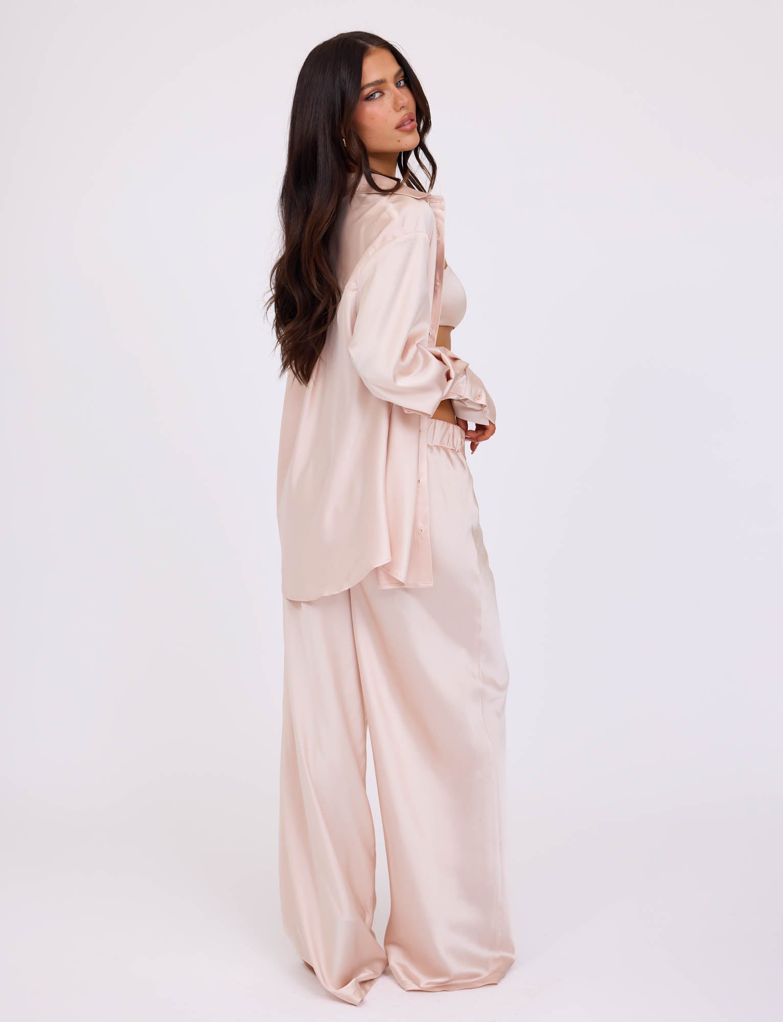 A woman with long dark hair stands in profile, dressed in a light pink, loose-fitting satin outfit from TIGERMIST. She wears the ALINA SHIRT - WHITE : IVORY, a long-sleeved button-up shirt over matching wide-leg pants. The plain white background emphasizes her stylish and relaxed ensemble.