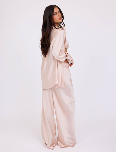 A woman with long dark hair stands in profile, dressed in a light pink, loose-fitting satin outfit from TIGERMIST. She wears the ALINA SHIRT - WHITE : IVORY, a long-sleeved button-up shirt over matching wide-leg pants. The plain white background emphasizes her stylish and relaxed ensemble.