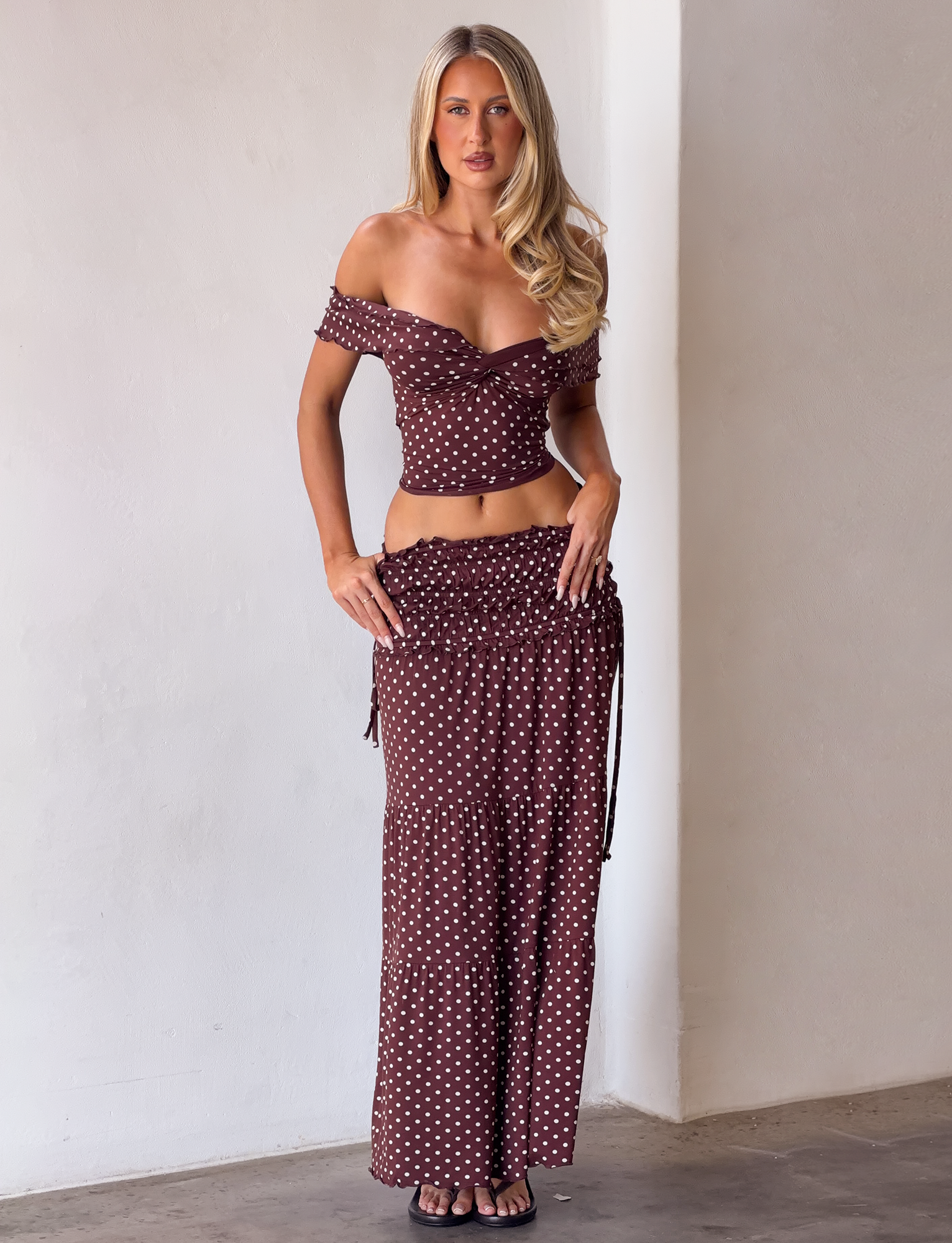 A woman stands indoors wearing the TIGERMIST DENALI TOP - BROWN DOT, an off-shoulder brown top with white polka dots and ruffled sleeves, paired with a matching long skirt and black sandals. She has long blonde hair and a neutral expression.