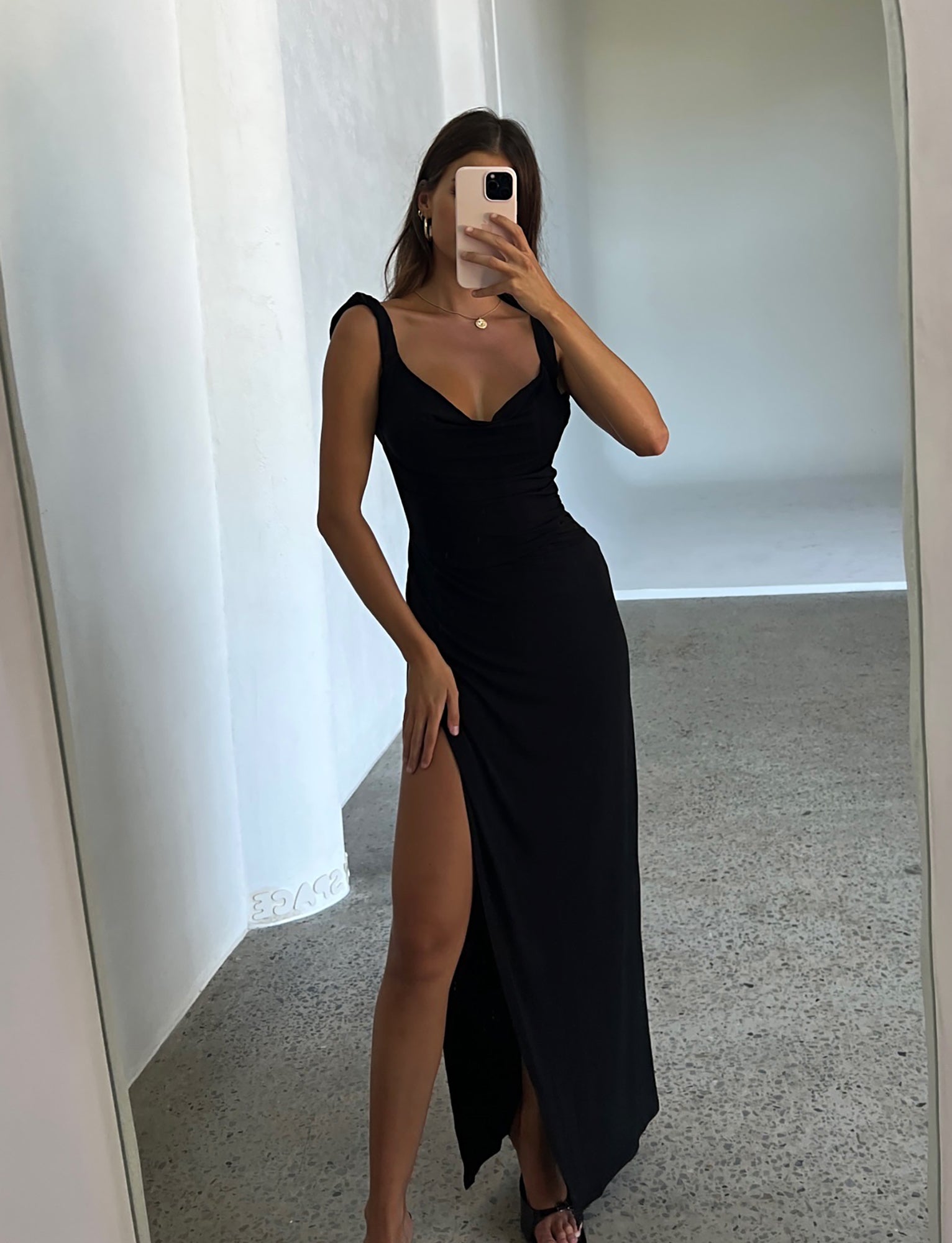 A woman is taking a mirror selfie in a minimalist room. She is wearing the JOSLIN MAXI DRESS - BLACK by TIGERMIST, which features a thigh-high slit, low neckline, and thin straps. She paired the dress with black heels. The room has light-colored walls and a polished stone floor.