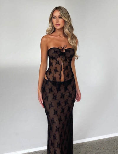 A woman with long blonde hair is standing against a plain white background. She is wearing the sophisticated KAROLINA SKIRT in black from TIGERMIST, featuring intricate lace patterns. She gazes slightly to the side with a serious expression, and her hands are relaxed by her sides.