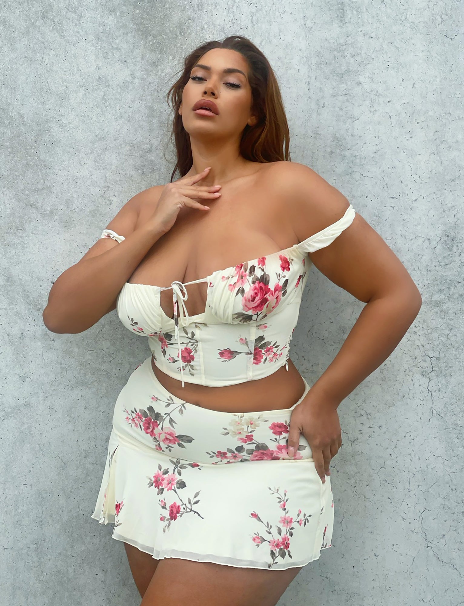 A woman stands confidently in front of a textured gray wall, wearing the TIGERMIST FEYRA TOP in yellow with a macro floral pattern. The top features off-the-shoulder sleeves and a small tie in the front. The matching short skirt is adorned with a similar pink and red floral design. She poses with one hand on her chest.