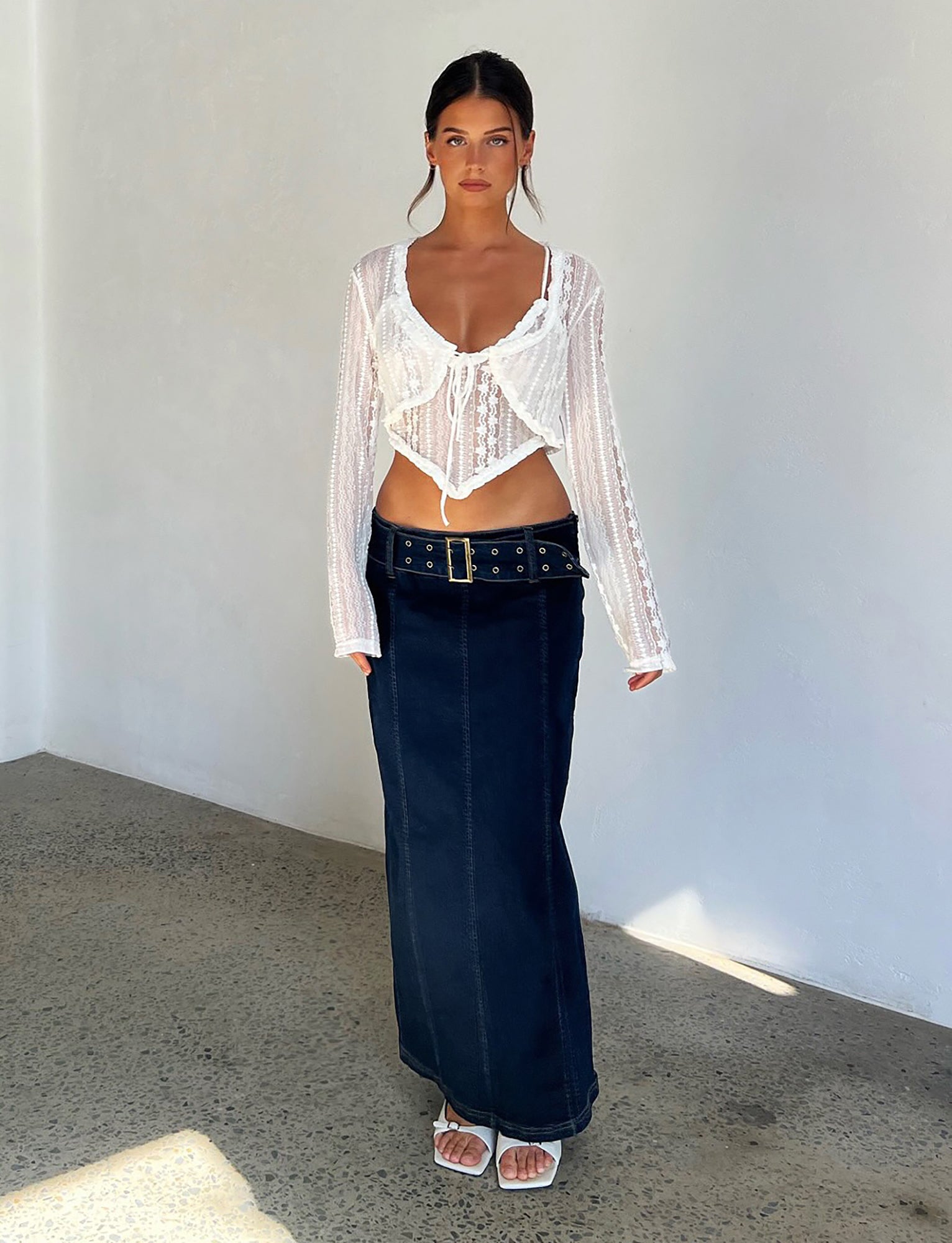 A woman stands in a minimalist room with a concrete floor and white walls. She is dressed in the SOPHA Two Piece Top by Tiger Mist, paired with a high-waisted dark blue denim maxi skirt featuring a wide belt, and white sandals. Her hair is styled in a neat updo, and she has a serious expression.