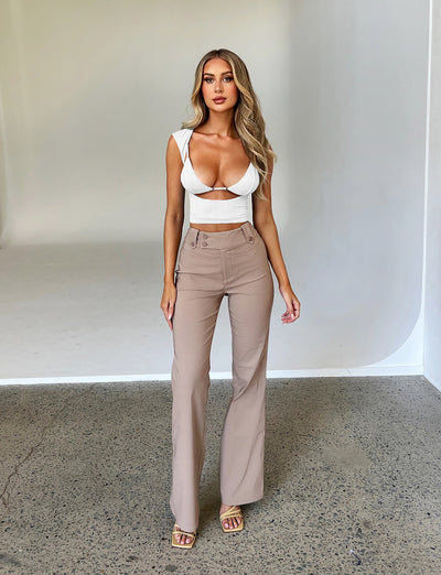 A woman with long hair stands posing against a plain backdrop. She is wearing a white, fitted crop top with cutouts and the KITTIE HIGH RISE PANT - BEIGE : TAN from TIGERMIST. She is also wearing beige open-toe sandals. Her expression is neutral, and her hands are relaxed by her sides.