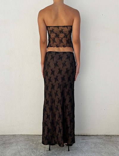 A person is standing with their back to the camera, wearing a strapless black mesh top and the Karolina Skirt from TigerMist. The outfit features a pattern of black floral designs. They are against a plain, light-colored wall and standing on a light-colored surface.
