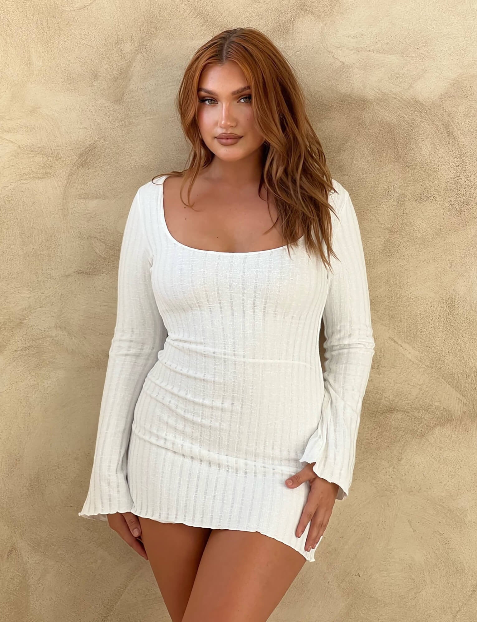 A woman with long, auburn hair poses against a beige textured background. She is wearing the Brenden Dress in a neutral color, a white, long-sleeved ribbed mini dress with a scoop neckline from TIGERMIST. She has a confident expression and is looking directly at the camera.