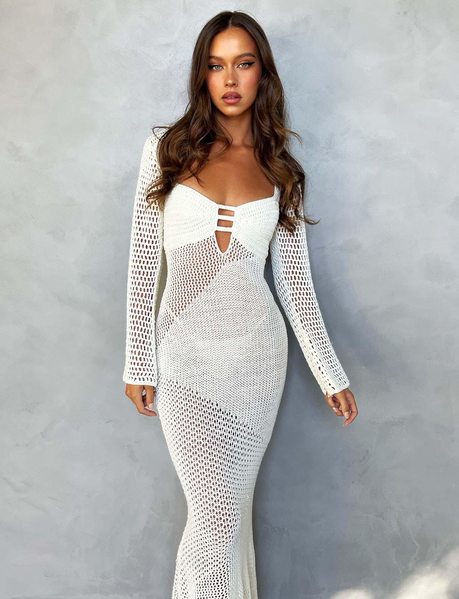 A woman with long brown hair poses against a gray background, wearing the MACAULEY DRESS - WHITE : CREAM by TIGERMIST. The dress is fitted, with long sleeves and geometric crochet patterns. She has a serious expression and her arms are relaxed by her sides.