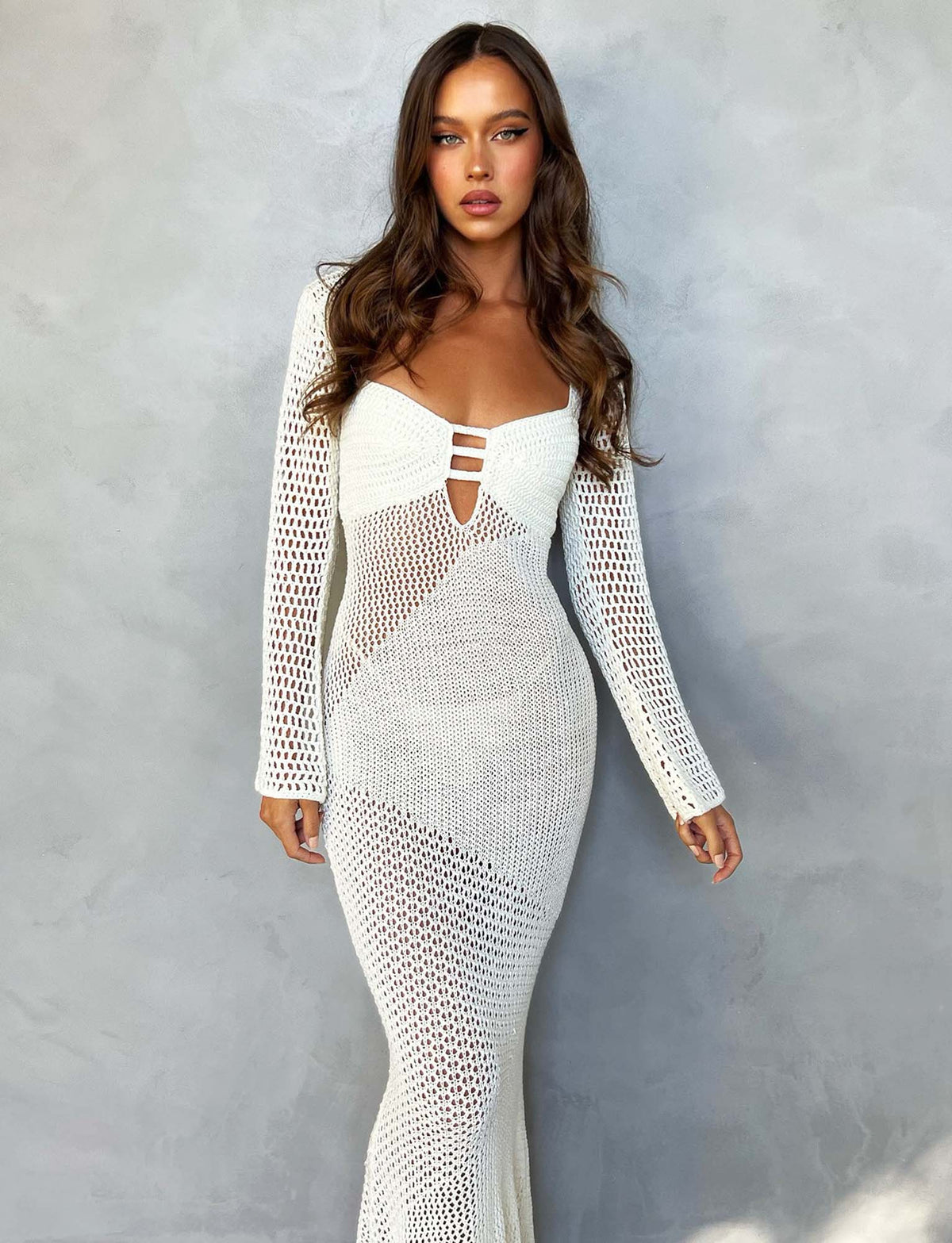 A woman with long brown hair poses against a gray background, wearing the MACAULEY DRESS - WHITE : CREAM by TIGERMIST. The dress is fitted, with long sleeves and geometric crochet patterns. She has a serious expression and her arms are relaxed by her sides.