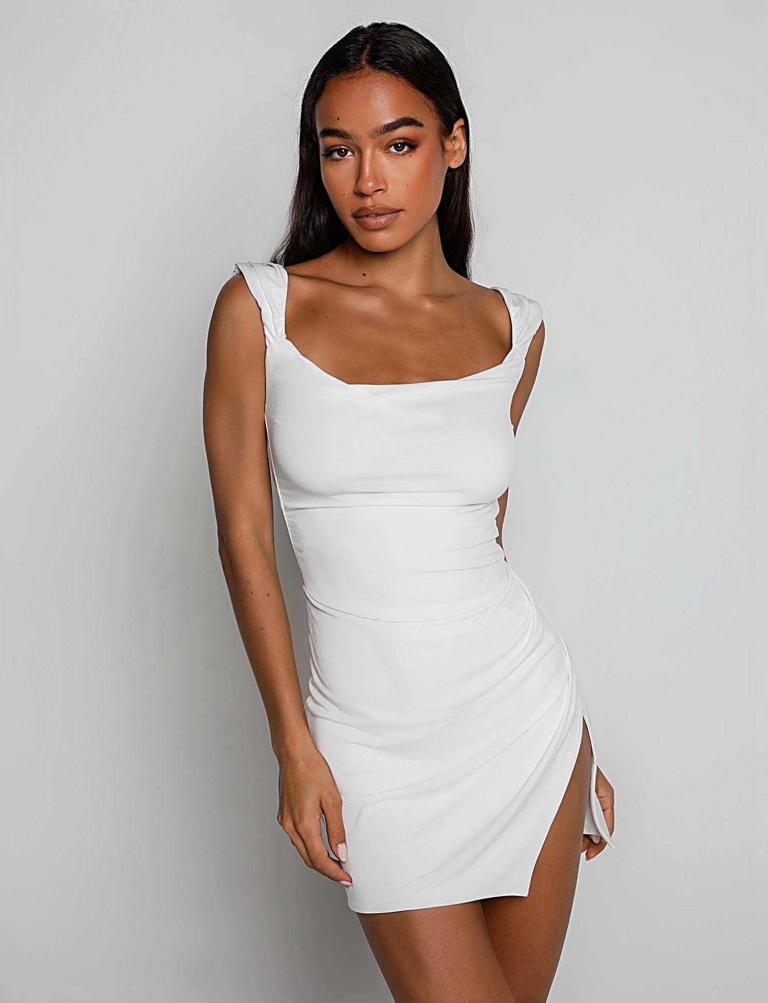 A woman with long dark hair is wearing the JOSLIN DRESS - WHITE from TIGERMIST. The form-fitting white sleeveless dress features a square neckline and a high slit on one side. She has a neutral expression and stands against a plain white background.