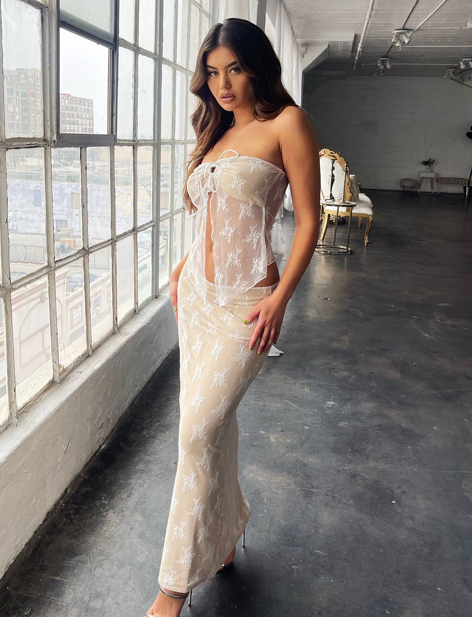 A woman wearing the Karolina Skirt in white lace from Tigermist confidently poses near large industrial-style windows in a spacious, minimalist room. Her long hair flows gracefully as she stands next to a golden chair and a table with a plant in the background.