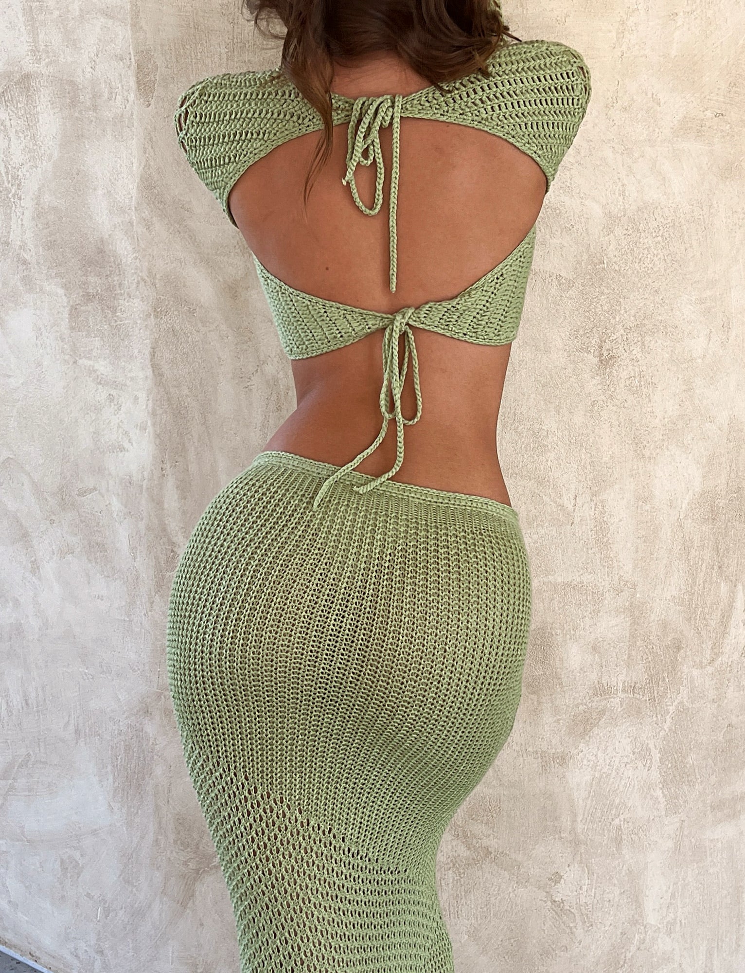 A person wearing the MACAULEY TOP - GREEN in pastel green, a backless, knitted top tied with a string, and a matching long, fitted skirt from TIGERMIST. Their back is visible as they stand against a textured beige wall, facing away from the camera.