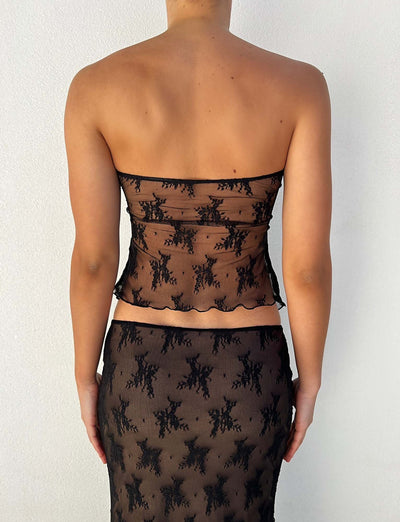 A person is shown from behind, wearing a stunning strapless KAROLINA TOP - BLACK and a matching semi-sheer black lace skirt from TIGERMIST. The background is a plain white wall.