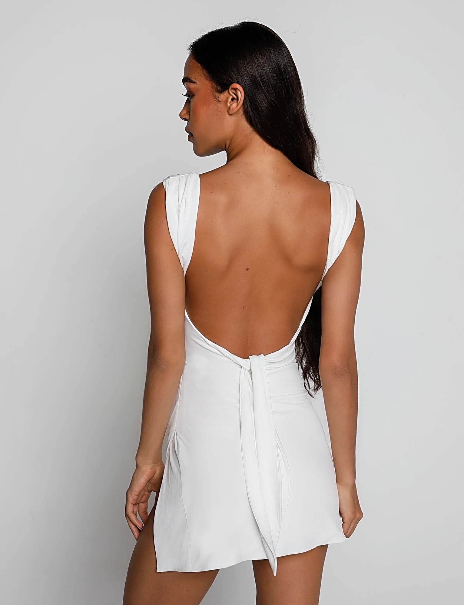 A woman with long, dark hair is seen from the back wearing the JOSLIN DRESS - WHITE by TIGERMIST. The short, white dress features wide straps and a backless design with a tie at the lower back, showcasing a flowy and elegant style. The background is plain white.