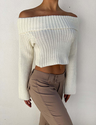 A person wearing the Tigermist ASHLYN JUMPER in white, an off-the-shoulder, long-sleeved knit sweater, paired with beige high-waisted pants stands against a plain wall. The outfit reveals the midriff and maintains a casual yet stylish appearance.