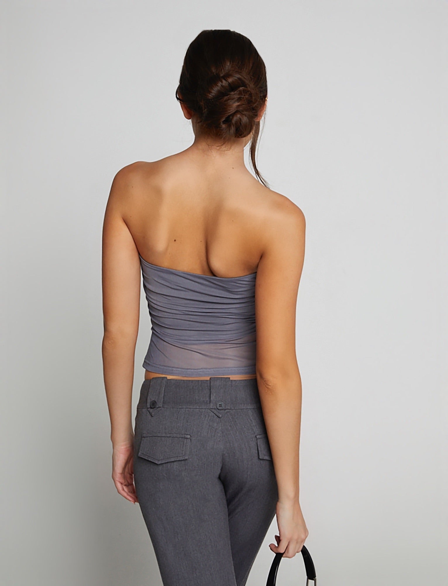 A woman with her back to the camera is wearing the HARRIETT TUBE TOP - GREY by TIGERMIST, paired with fitted gray pants. She holds a black handbag in her right hand, and her hair is elegantly styled in a low bun.
