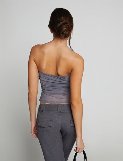 A woman with her back to the camera is wearing the HARRIETT TUBE TOP - GREY by TIGERMIST, paired with fitted gray pants. She holds a black handbag in her right hand, and her hair is elegantly styled in a low bun.