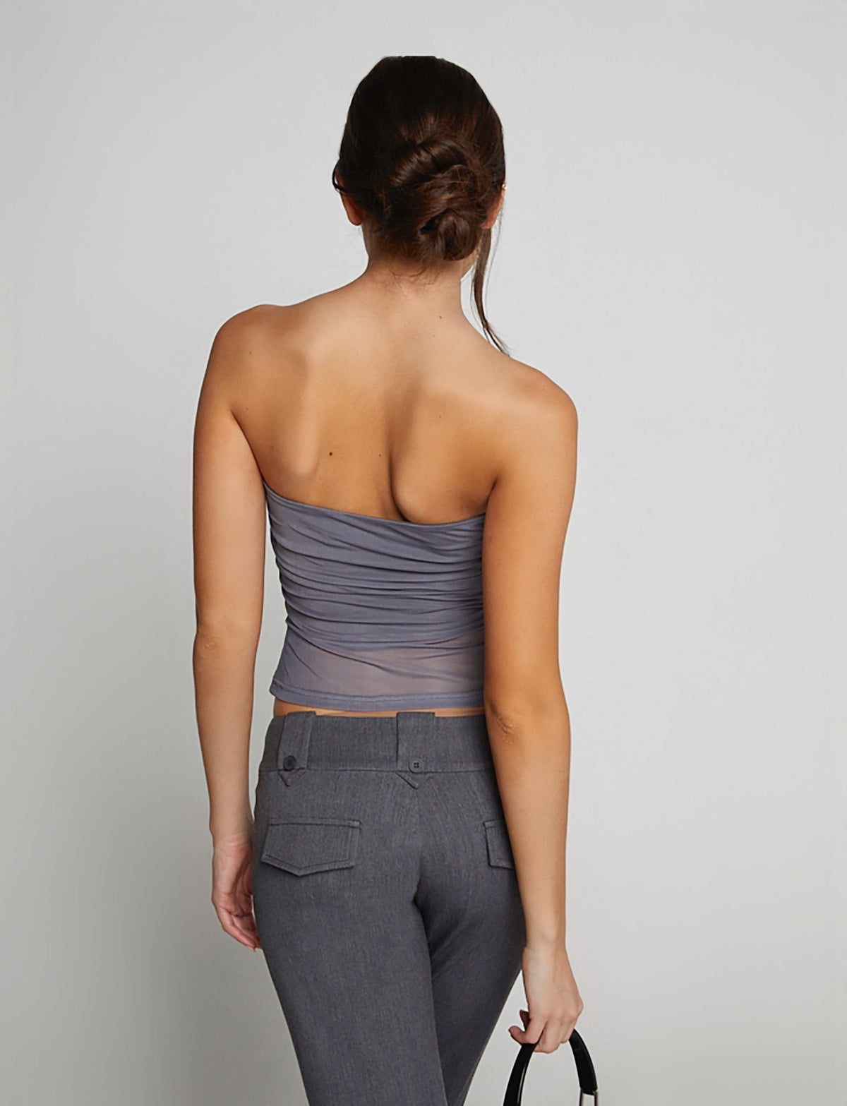 A woman with her back to the camera is wearing the HARRIETT TUBE TOP - GREY by TIGERMIST, paired with fitted gray pants. She holds a black handbag in her right hand, and her hair is elegantly styled in a low bun.