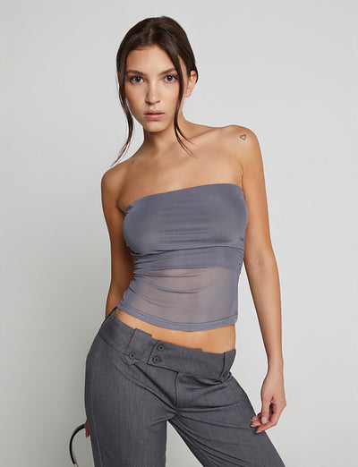 A woman stands against a plain background wearing the HARRIETT TUBE TOP - GREY from TIGERMIST and gray trousers with button details. She has her hair pulled back, with loose strands framing her face and a small heart tattoo on her left shoulder.