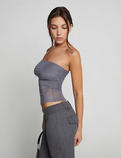 A woman with long hair, wearing the HARRIETT TUBE TOP – GREY from TIGERMIST along with high-waisted grey pants, stands slightly turned to the side and looks downward against a plain white background. She holds a small black bag in her left hand.