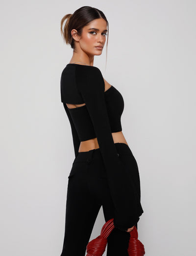 A woman stands against a plain background, posing sideways in a sleek, modern outfit from TIGERMIST. She wears the SOPH TWO PIECE TOP - BLACK with cut-out details at the back and matching black pants. Holding a red, coiled accessory in one hand and with her hair tied back, she exudes contemporary style.