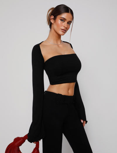 A person with long hair tied back in a ponytail is standing against a plain background. They are wearing the chic SOPH TWO PIECE TOP - BLACK and matching black pants from TIGERMIST, holding a red textured purse in their left hand.