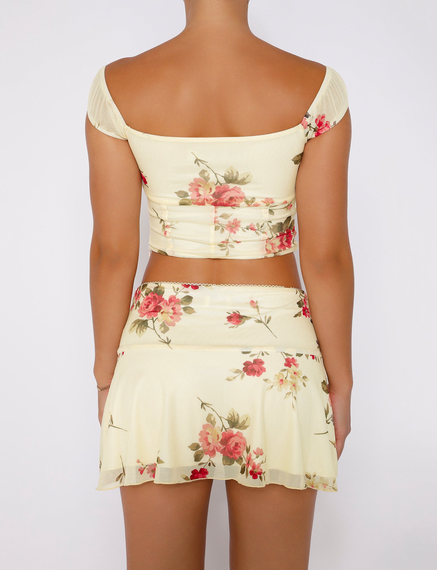 Rear view of a person wearing the FEYRA SKIRT - YELLOW : MACRO FLORAL : POP FLORAL matching two-piece outfit from TIGERMIST with a floral pattern. The outfit consists of an off-the-shoulder, cropped top and a short A-line skirt, both in light cream with red and pink roses. The background is plain white.