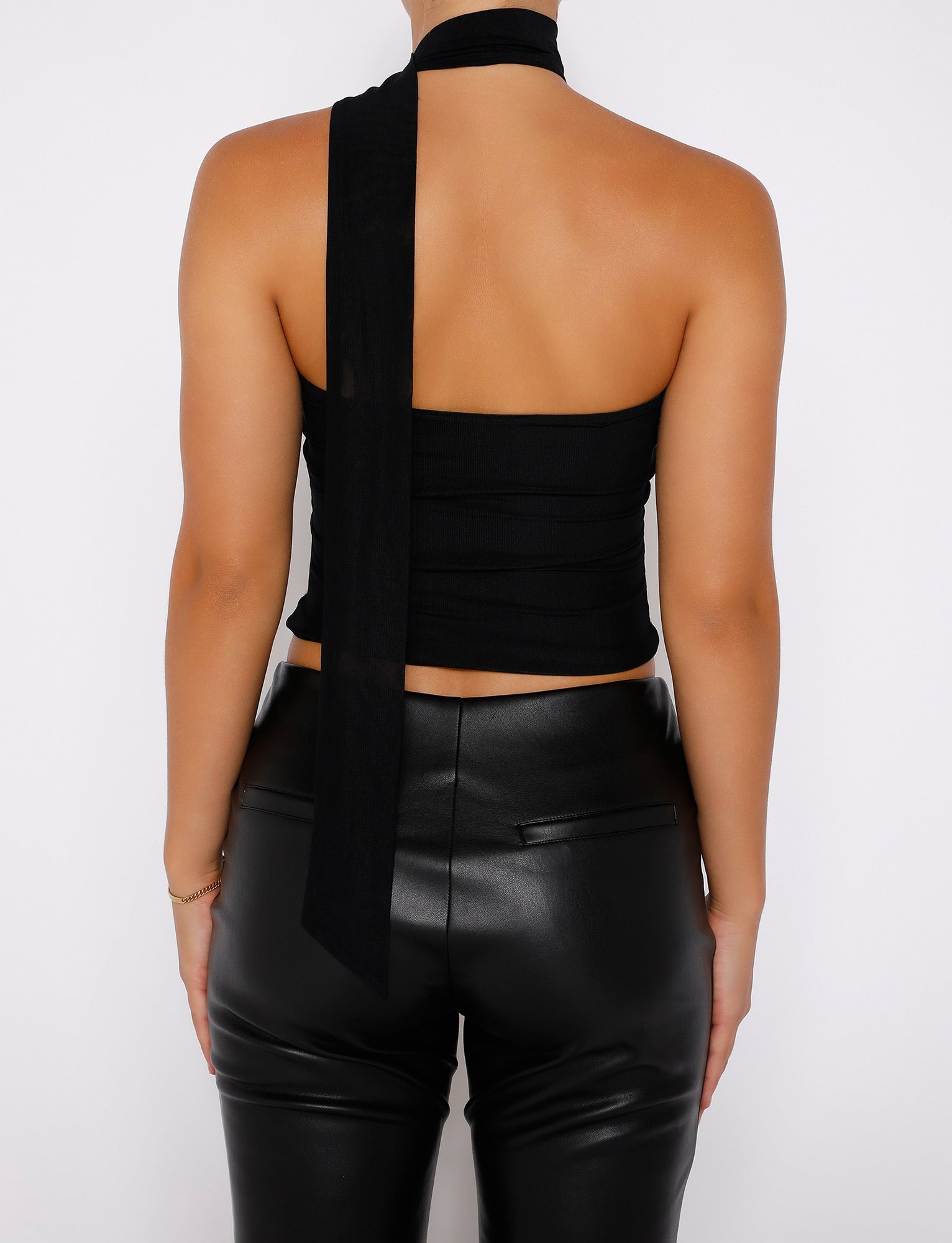A person stands with their back to the camera, wearing a TIGERMIST CHRYSLER TOP in black, featuring a long ribbon tie and paired with black leather pants. The halter neck design wraps around the neck and extends down the back, while the person has an arm clasped behind them.