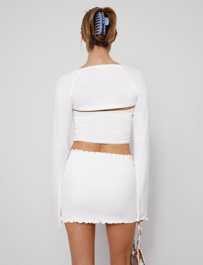 A person stands against a plain background, facing away from the camera. They are wearing the SOPH TWO PIECE TOP - WHITE, which features a long-sleeve cropped top and a matching white mini skirt with scalloped edges from TIGERMIST. Their hair is tied up with a blue hair clip, and they are holding a colorful accessory.