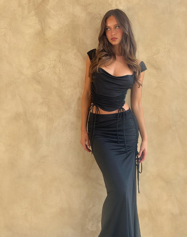 A woman with long wavy hair poses against a beige textured wall, wearing a fitted black dress with ruched details and side ties.