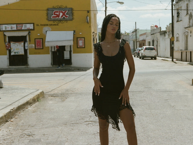 A woman in a black lace dress stands confidently in the middle of a sunny street near a yellow building with a "SIX" sign, surrounded by old white buildings and parked cars.