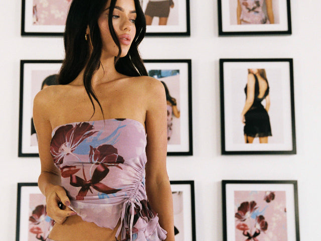 A woman in a pink floral strapless top stands in front of a wall decorated with framed fashion photos, some featuring similar floral patterns as her top.