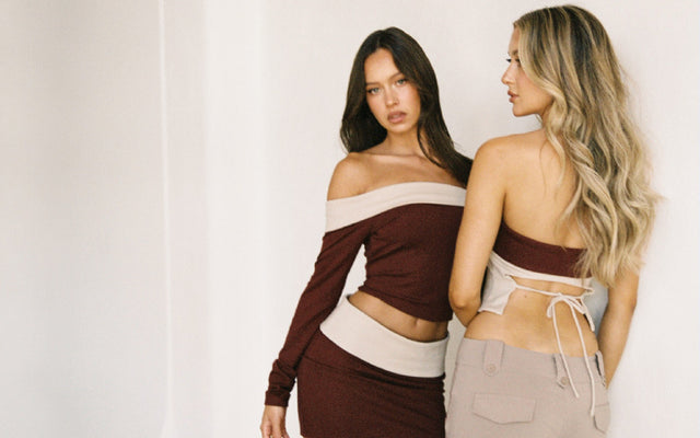 Two women pose together against a white backdrop. One wears a maroon off-shoulder top and matching skirt, while the other wears a beige backless top with beige pants. Both have long, loose hair.