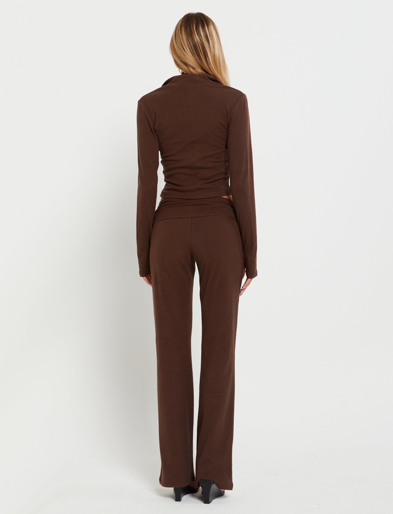 A woman with long, straight hair stands facing away from the camera. She is wearing a brown, long-sleeved, form-fitting Kavat Jacket from Tiger Mist along with matching brown pants. The outfit appears to be made of a comfortable, stretchy fabric, and she is wearing black heeled shoes. The background is plain white.