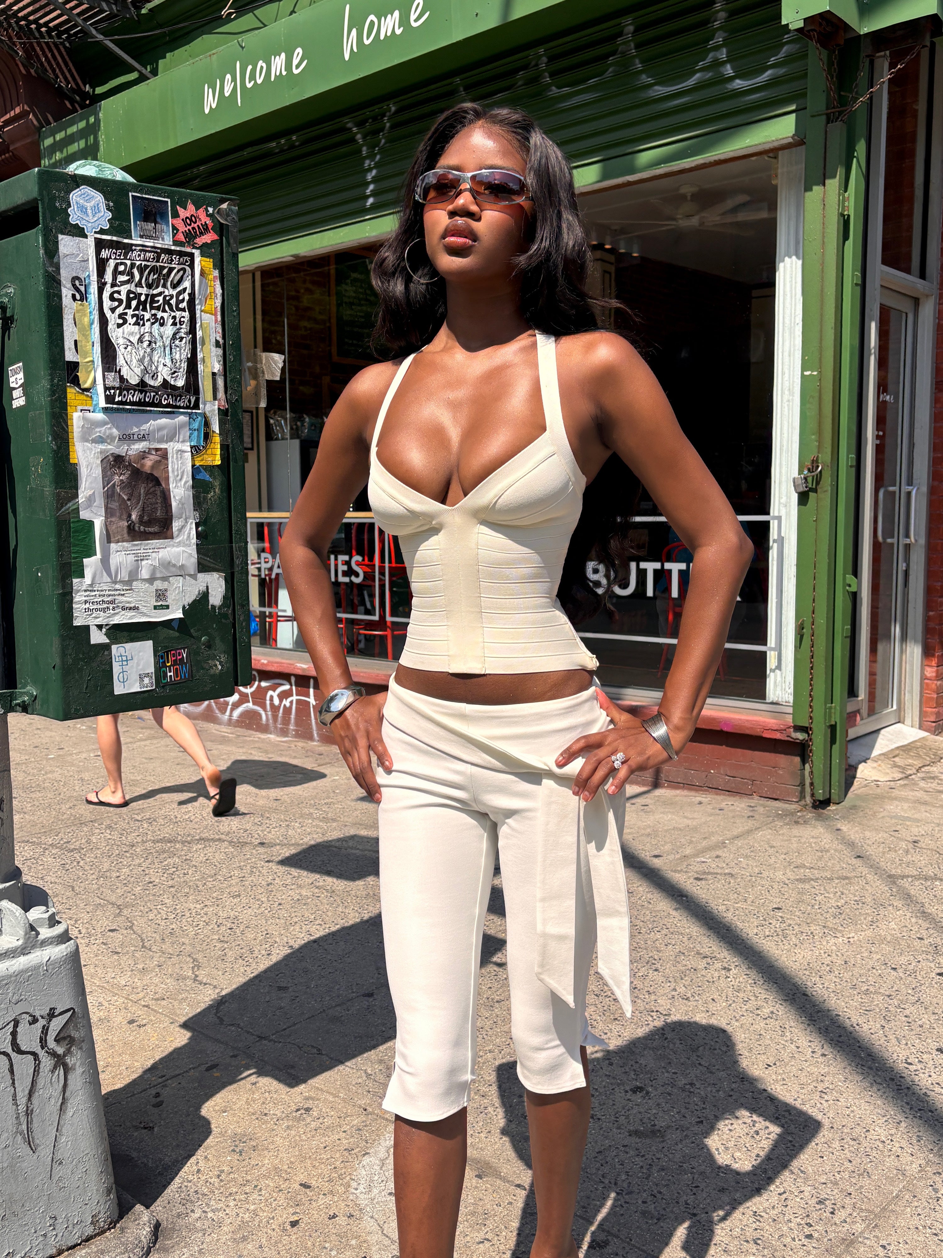 A woman in the SYDEL BANDAGE TOP - CREAM by TIGERMIST and sunglasses stands confidently on a sunny city sidewalk in front of a storefront with green signage reading “Welcome Home.”.