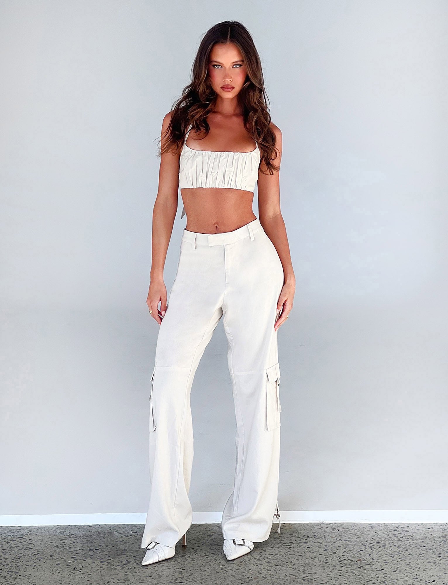 A woman is standing against a plain gray background, adorned in a white, ruched crop top and high-waisted KAYA PANT - WHITE : CREAM from TIGERMIST. She completes her look with white pointed-toe shoes. With her arms relaxed by her sides, she gazes directly at the camera with a neutral expression.