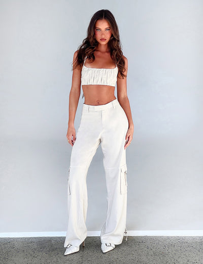 A woman is standing against a plain gray background, adorned in a white, ruched crop top and high-waisted KAYA PANT - WHITE : CREAM from TIGERMIST. She completes her look with white pointed-toe shoes. With her arms relaxed by her sides, she gazes directly at the camera with a neutral expression.