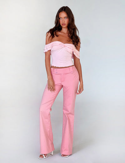 A woman stands against a plain light background, wearing a light pink off-the-shoulder top adorned with ruffle details and the KITTIE PANT in pink from TIGERMIST. She complements her outfit with white high-heeled sandals. Her long, wavy brown hair flows freely as she looks towards the camera with a composed expression.