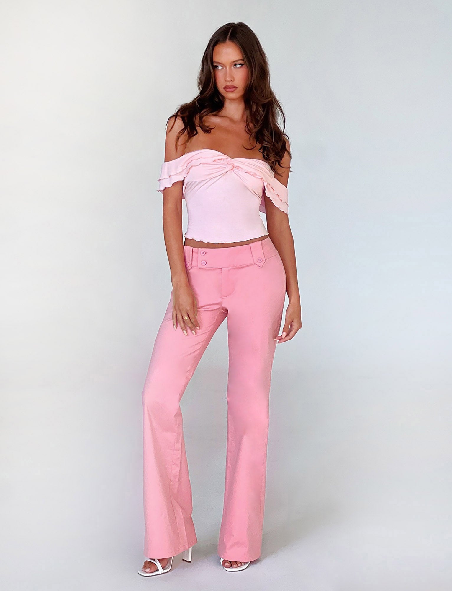 A woman stands against a plain backdrop wearing the DENALI TOP - PINK and matching pink high-waisted pants from TIGERMIST. She has long brown hair and wears white open-toe high heels. Her left hand rests on her hip, and she gazes directly at the camera.