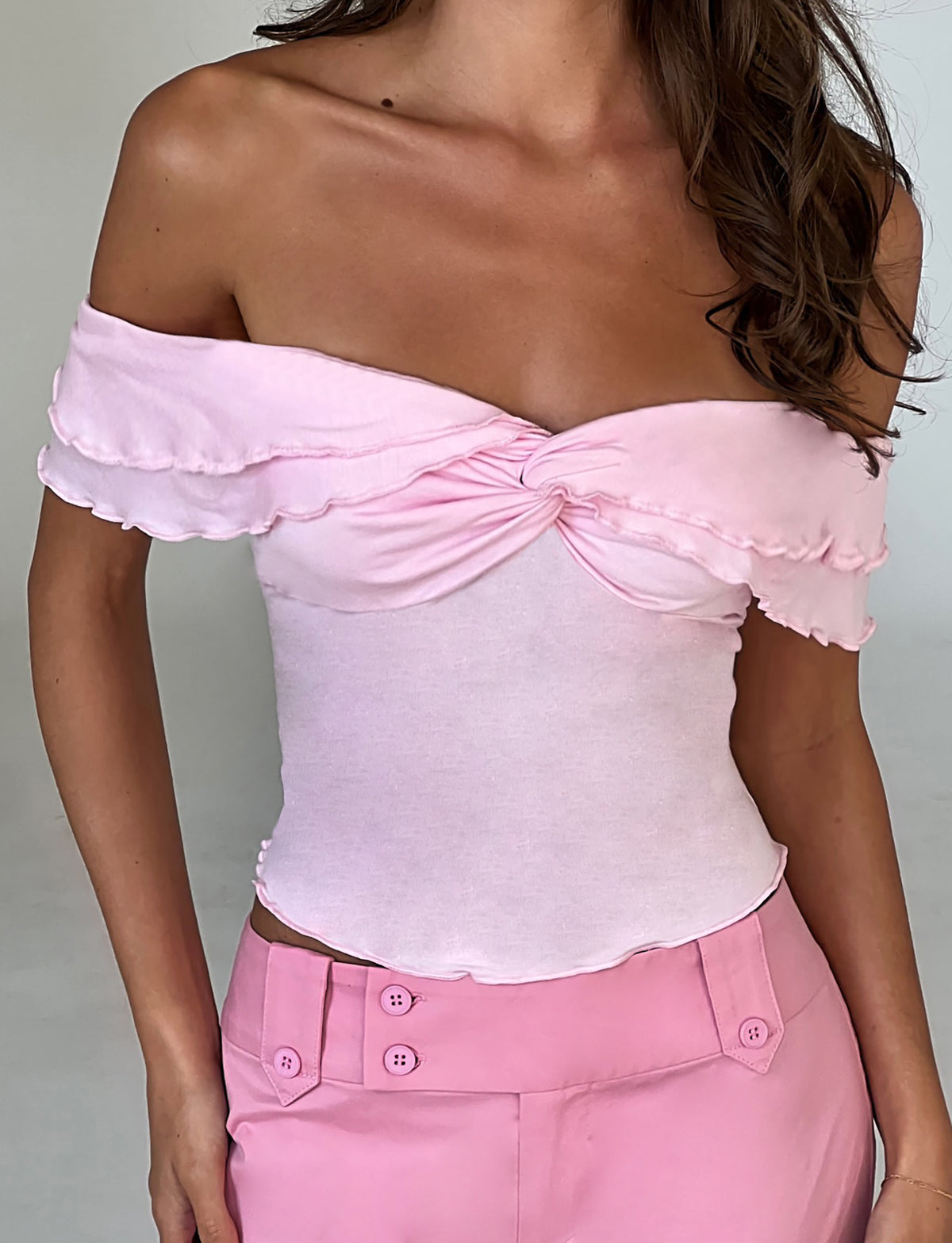 A woman is wearing the DENALI TOP - PINK by TIGERMIST, which features an off-the-shoulder design with ruffled edges along the neckline and sleeves, as well as a twist knot at the center of the bust. She pairs it perfectly with high-waisted pink pants that have a double button detail.
