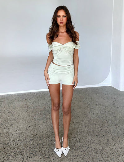 A woman with long, wavy brown hair stands on a textured concrete floor in a brightly lit room, showcasing her chic Tiger Mist off-shoulder cropped top and matching high-waisted Denali shorts in sage green, paired perfectly with white pointed high heels.
