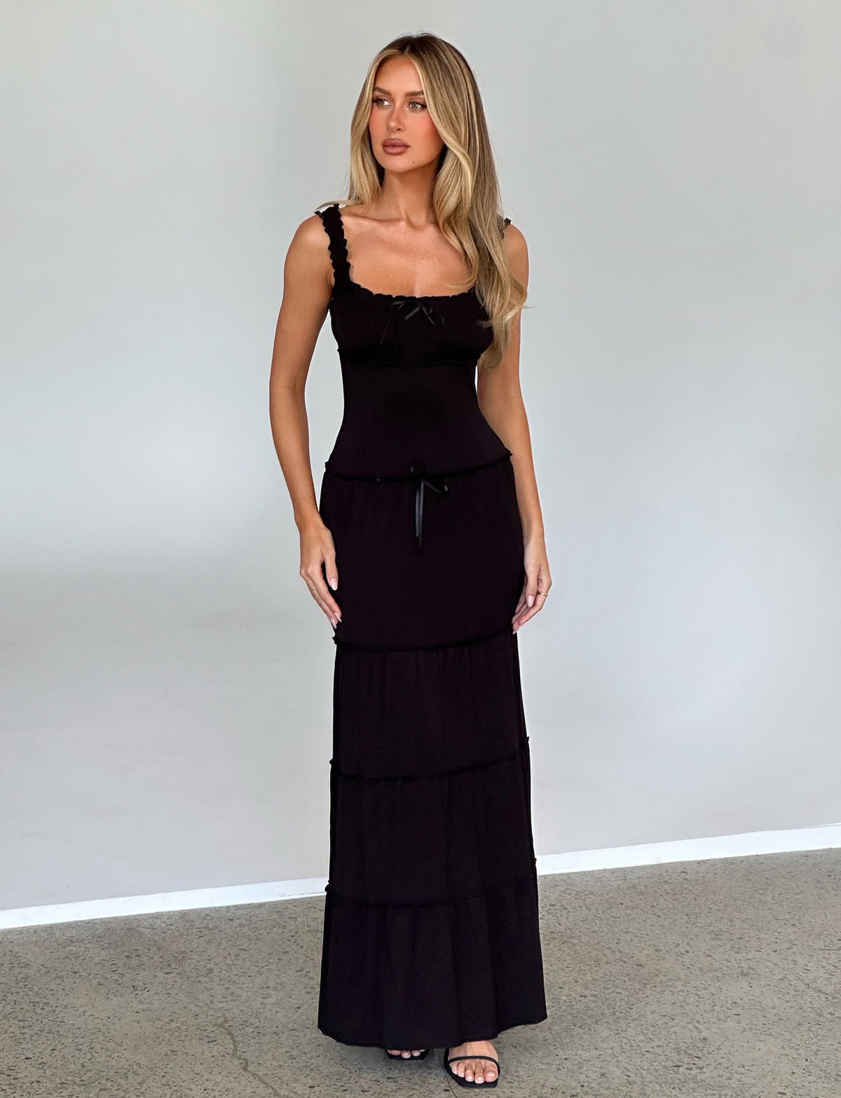 A woman with long blonde hair stands against a light gray background, wearing the NINA DRESS - BLACK from TIGERMIST. She has a neutral expression and her hands are relaxed by her sides. She is also wearing black sandals.