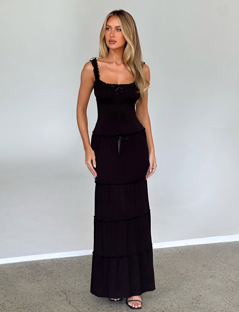 A woman with long blonde hair stands against a light gray background, wearing the NINA DRESS - BLACK from TIGERMIST. She has a neutral expression and her hands are relaxed by her sides. She is also wearing black sandals.