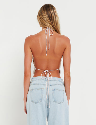 A woman with long blonde hair, wearing a TIGERMIST ASTRAEA TOP in gold and rose gold sequin, and light blue denim jeans is standing with her back to the camera. The top is tied at the neck and lower back, and the jeans have large pockets. The background is plain white.