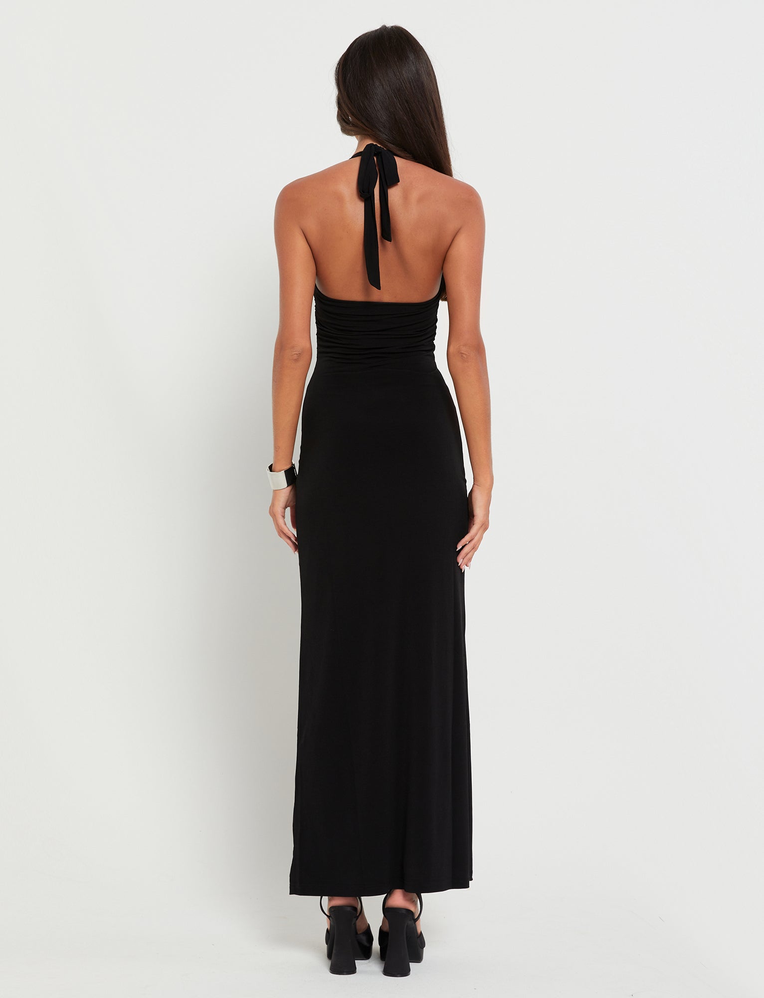 A woman with long, dark hair is wearing the CORALINE DRESS - BLACK by TIGERMIST. The dress is backless, floor-length, and ties around her neck. As she stands facing away from the camera, she showcases the back of the dress and her black high-heeled shoes against a plain white background.