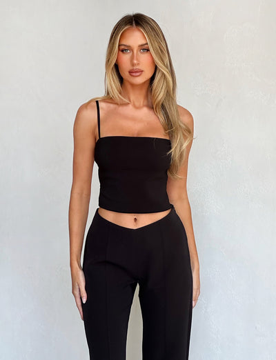 A woman with long blonde hair is posing against a light grey background. She is wearing the NAYSHA TOP - BLACK from TIGERMIST, a sleeveless, form-fitting top with thin straps, and matching high-waisted black pants. Her expression is neutral, and she has one arm relaxed by her side while the other is bent slightly.