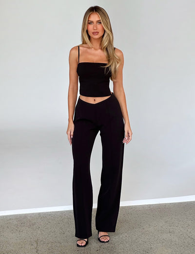 A person with long blonde hair is standing and posing against a plain background. They are wearing the NAYSHA TOP in black from TIGERMIST, black high-waisted trousers, and black open-toe heeled sandals. Their expression is neutral, and their posture is upright.