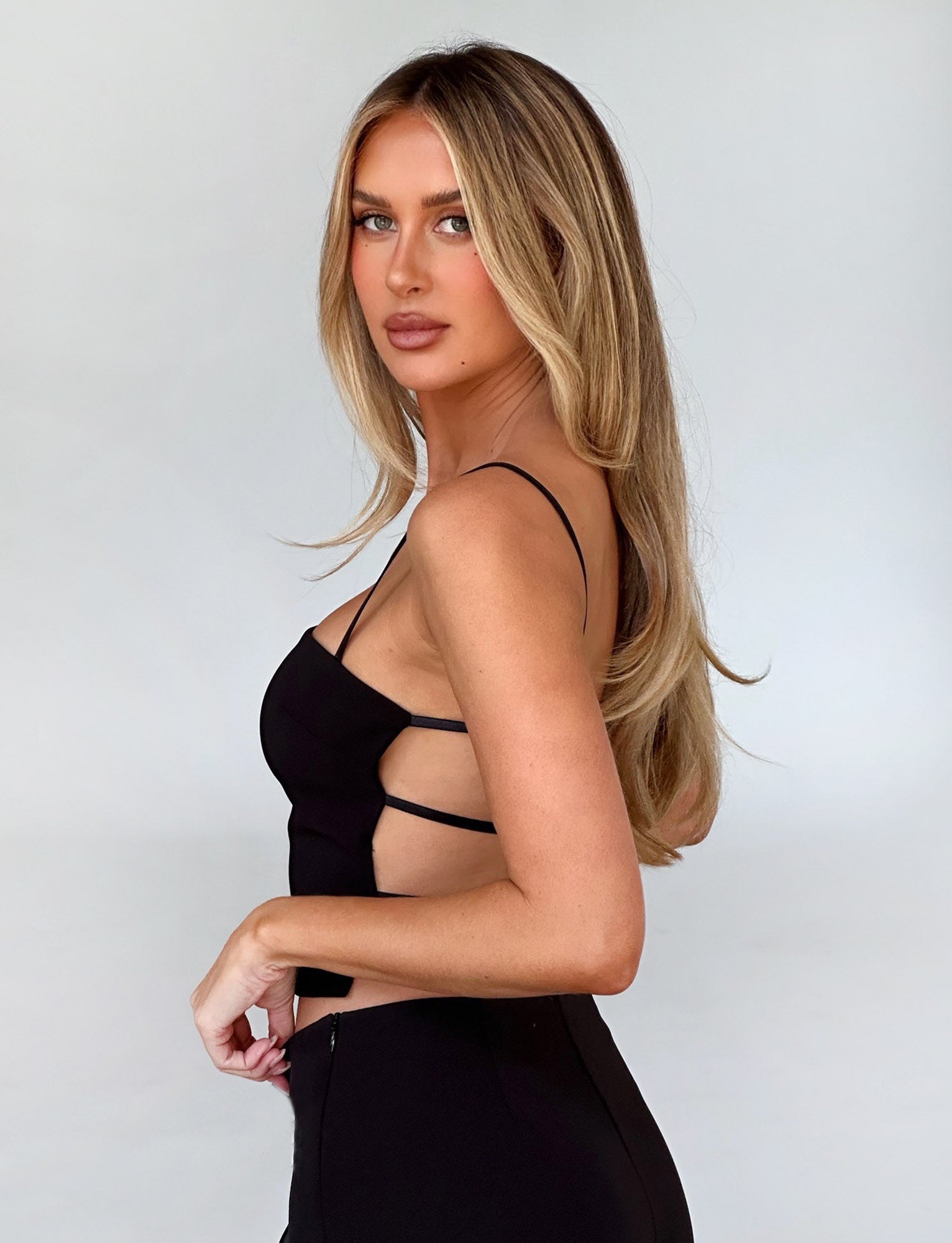 A woman with long blonde hair stands sideways, posing in the black NAYSHA Top by TIGERMIST against a plain, light background. She looks towards the camera with a calm expression, and her right hand rests on her hip.