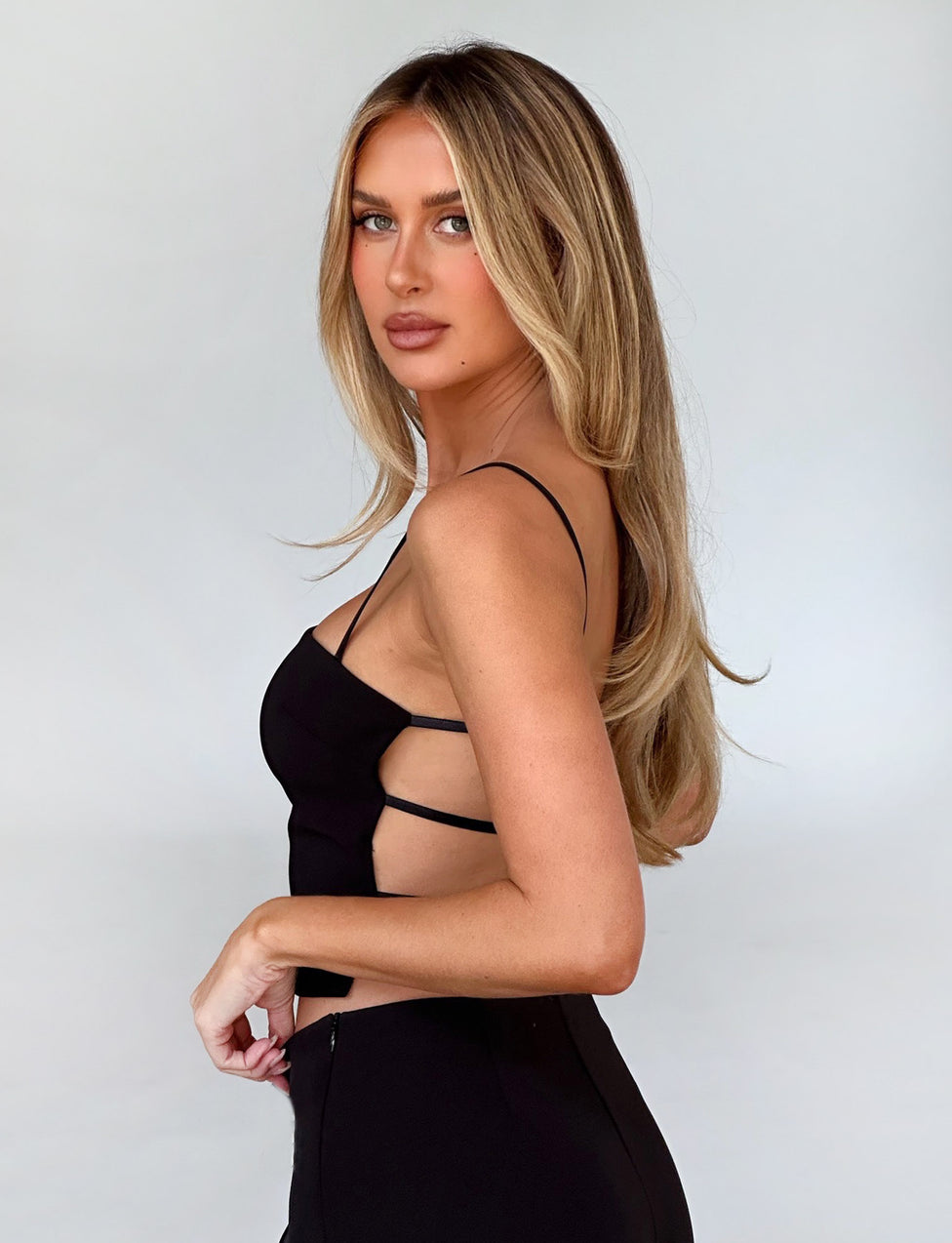A woman with long blonde hair stands sideways, posing in the black NAYSHA Top by TIGERMIST against a plain, light background. She looks towards the camera with a calm expression, and her right hand rests on her hip.