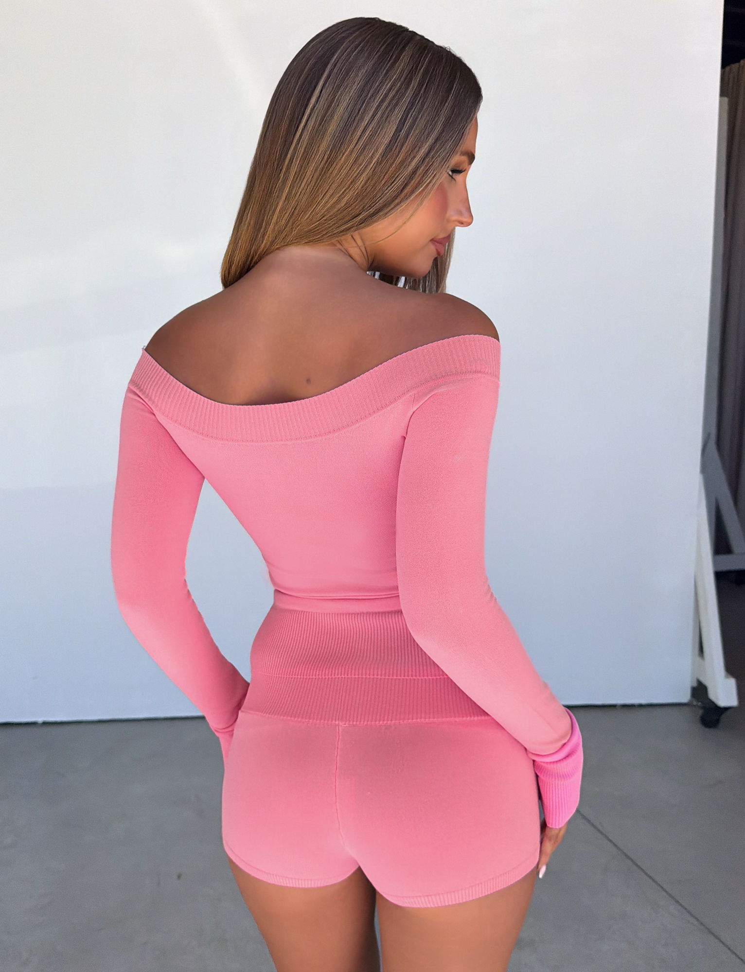 A woman with long hair poses with her back to the camera, wearing a fitted pink off-the-shoulder top paired with the TIGERMIST PARKER KNIT MICRO SHORT in coral, standing indoors on a concrete floor against a white wall.
