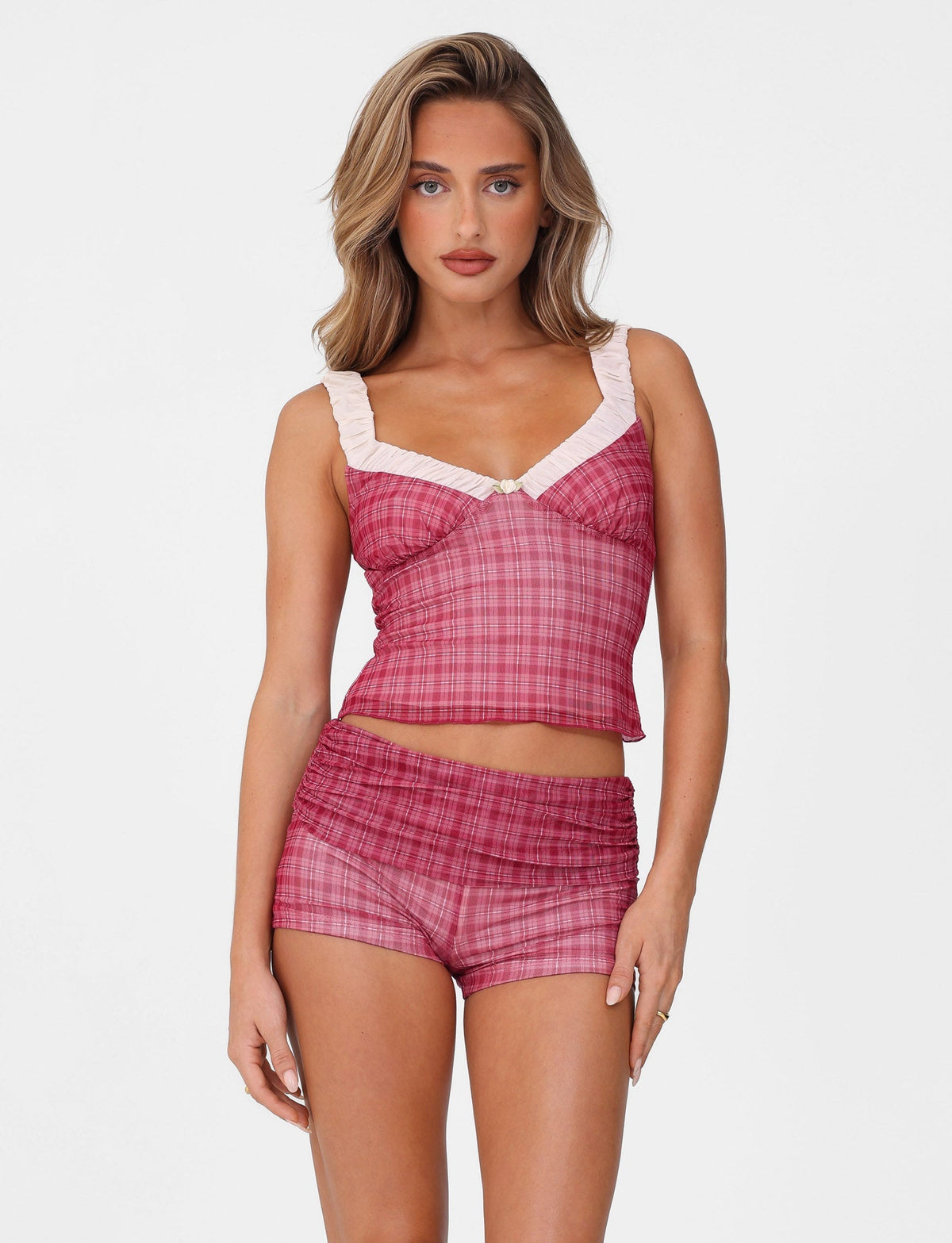 A person confidently wears the TIGERMIST AMARAN TOP - RED, a chic red check crop top with ruffled straps and matching shorts, styled like a bustier. Accented with light pink at the neckline, it stands out against a plain white backdrop.